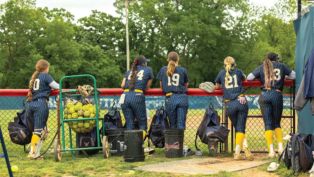 Softball students posing at the field, leaning against a fence with their backs turned to the camera